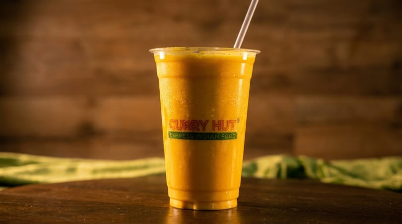 A Curry Hut mango lassi in a clear plastic cup with a straw on a wooden table.
