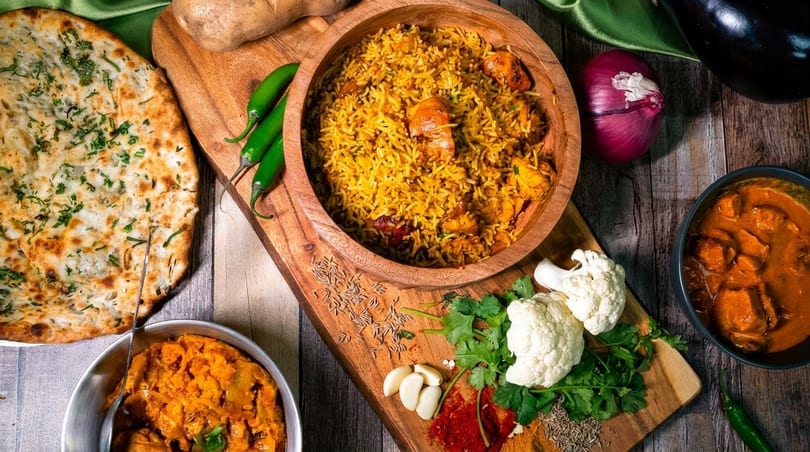 An overhead shot of Indian dishes including biryani, naan, curry, and fresh spices on a wooden table.