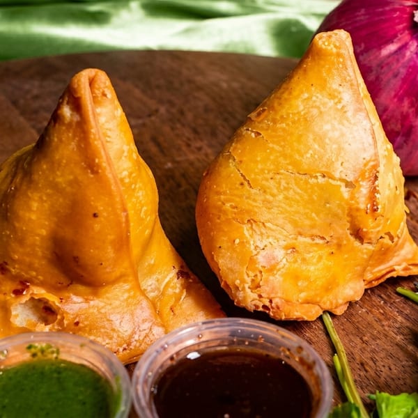 Two golden-brown samosas with green and brown dipping sauces, cilantro, and a red onion on a wooden board.