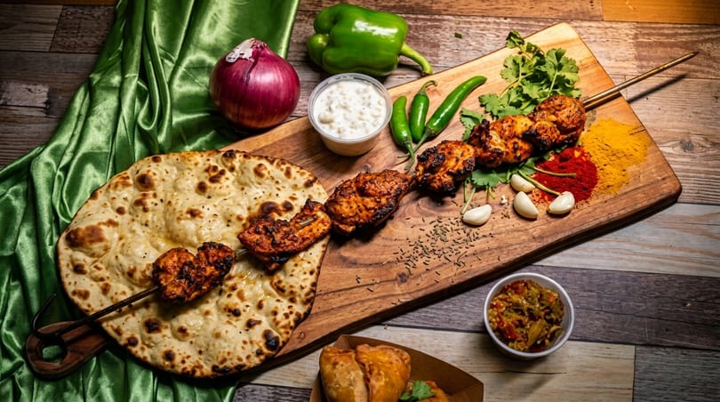 Grilled chicken skewers, naan bread, spices, and fresh vegetables on a wooden board.