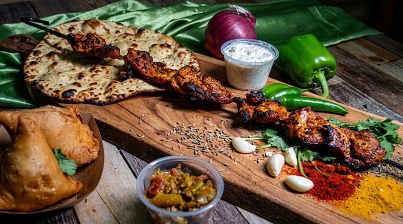 Grilled chicken skewers, naan bread, samosas, spices, and vegetables on a wooden board.