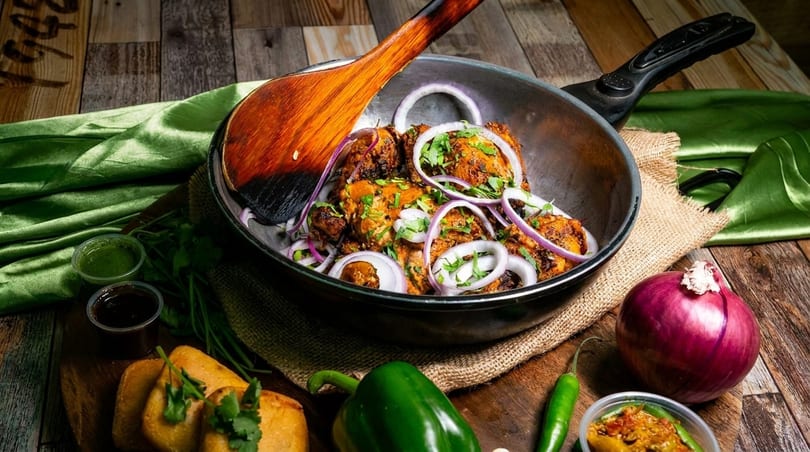 Chicken tikka in a pan with red onions and cilantro, surrounded by spices and sauces.