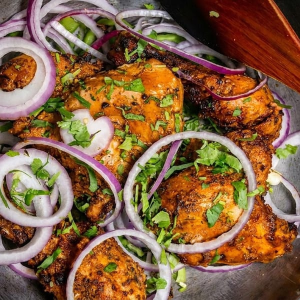Spicy chicken tikka pieces garnished with red onion rings and fresh cilantro in a metal bowl.