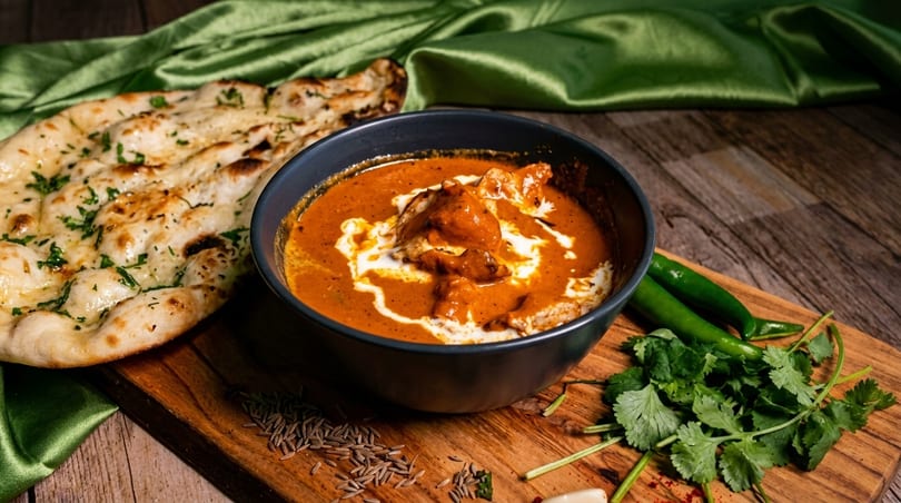 Butter chicken in a black bowl with naan bread, green chilies, and cilantro on a wooden board.