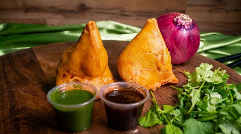 Two golden-brown samosas with green and brown dipping sauces, a red onion, and fresh cilantro.