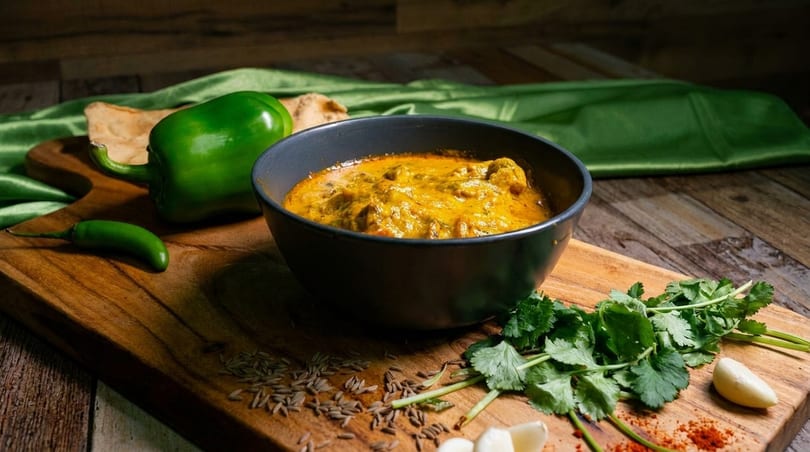 A bowl of yellow curry with green peppers, cilantro, and spices on a wooden board.