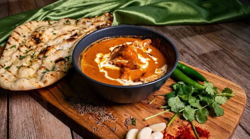 Butter chicken with naan bread, cilantro, green chilies, and spices on a wooden board.